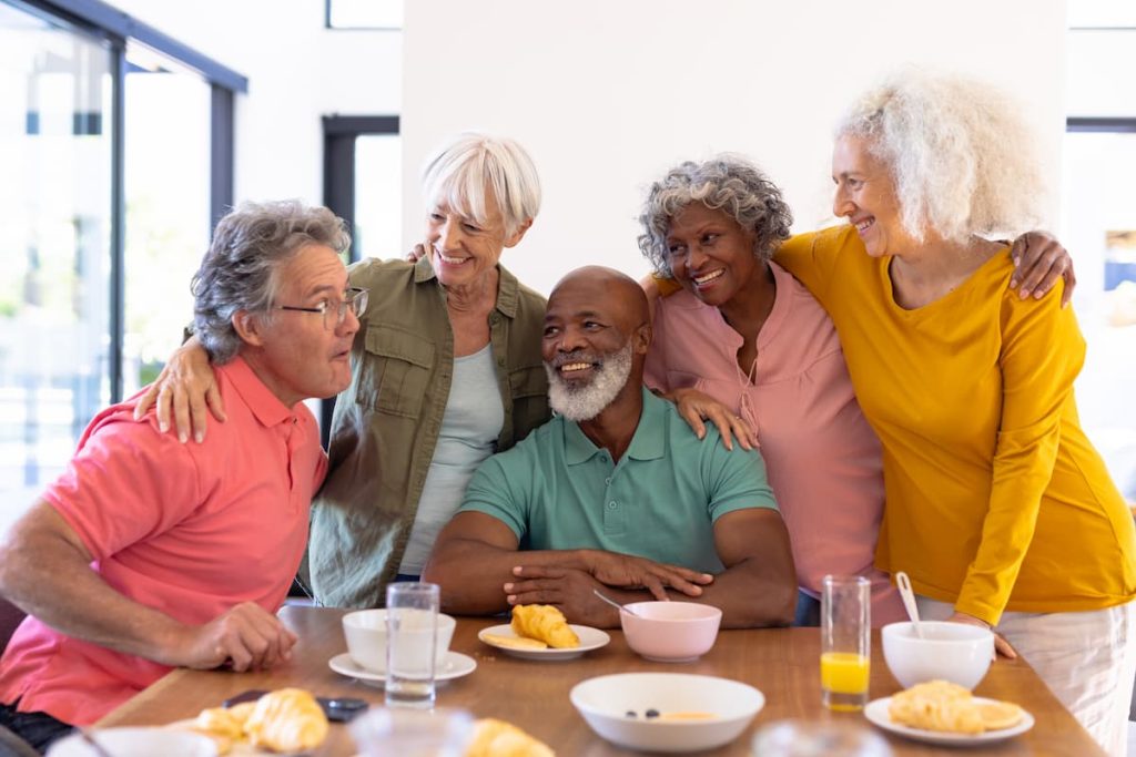 multiracial-senior-friends-with-arms-around-enjoying-by-breakfast-dining-table-nursing-home-cheerful-unaltered-food-drink-healthy-togetherness-support-assisted-living-retirement