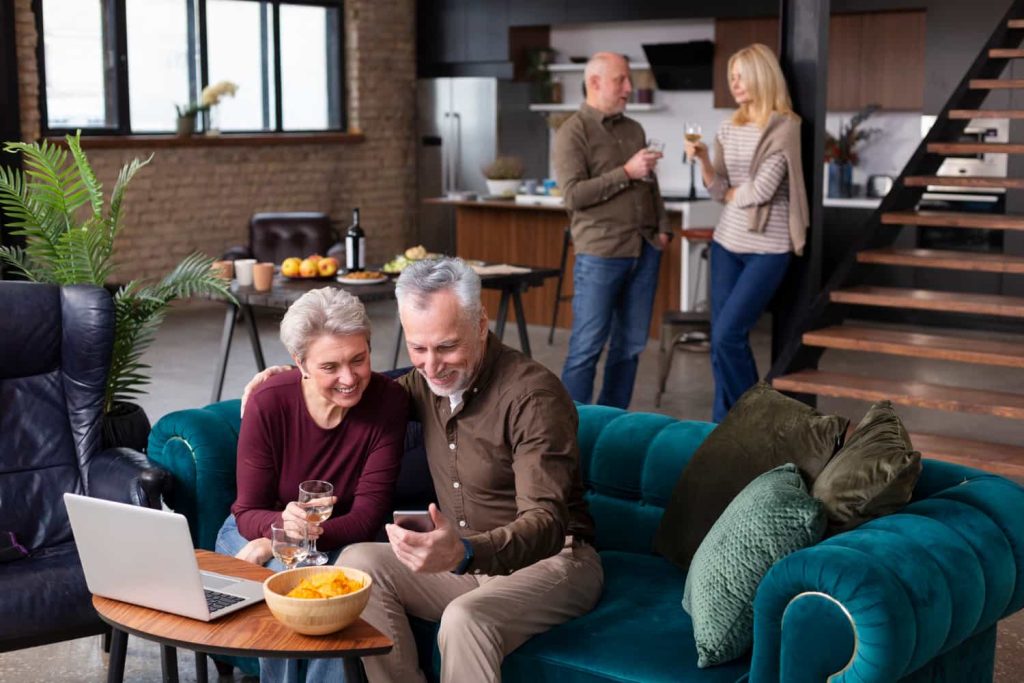 Older couple smiling on a teal sofa while looking at a phone, symbolizing inclusion, connection, and supportive home sharing in Vancouver.