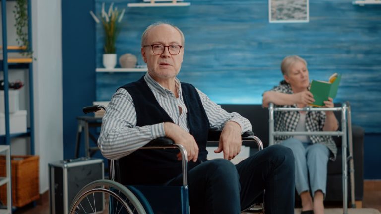 Senior man in a wheelchair sitting in a bright living room with another person reading in the background, representing supportive housing and independent living in Vancouver.